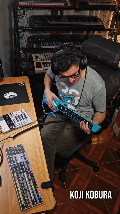 Person sitting at a desk with electronic equipment, wearing headphones and holding a Cyber-G guitar.