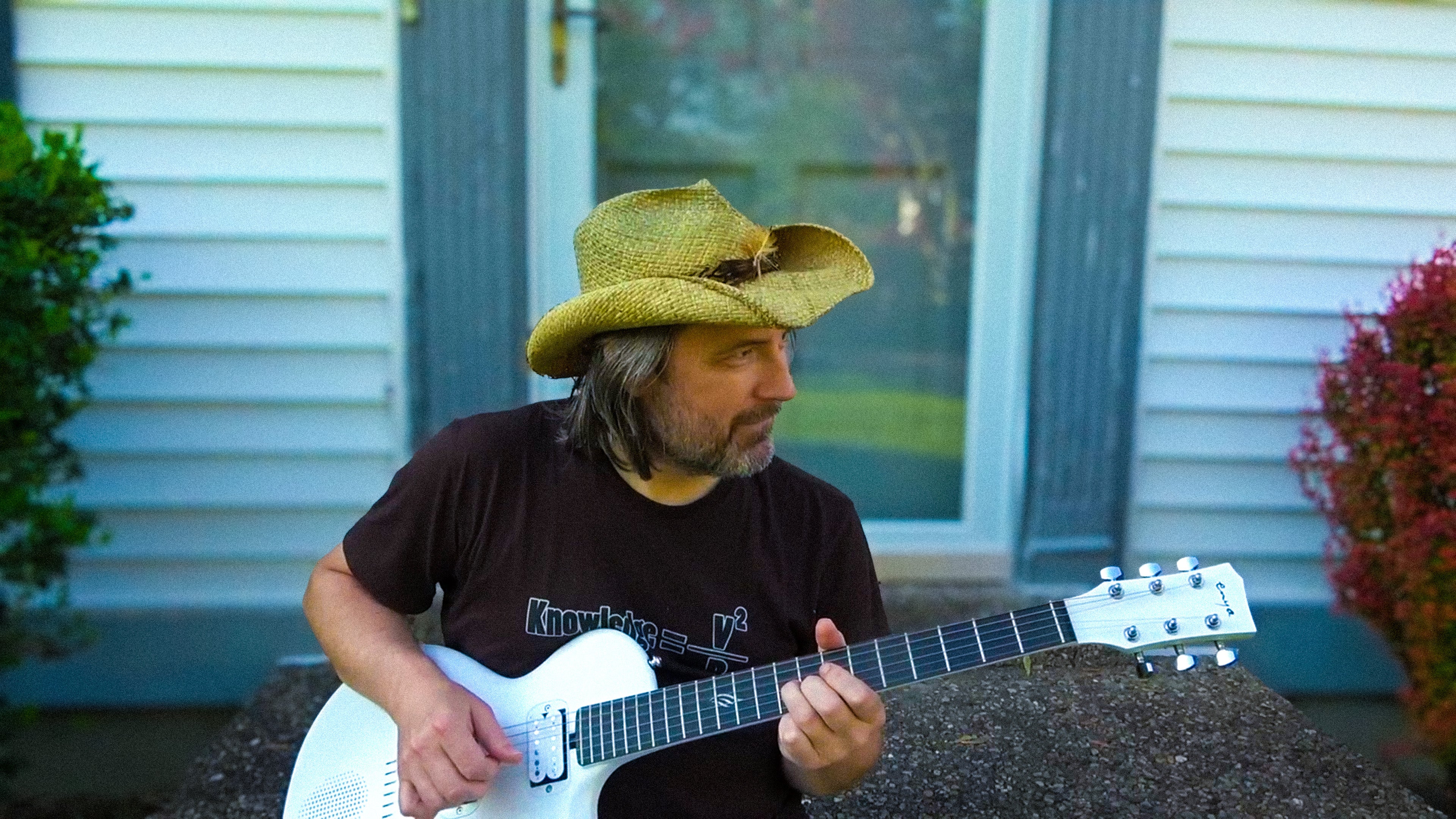 The Guitologist Brad sitting on the ground in front of door and playing a pearl white Nova Go Sonic.